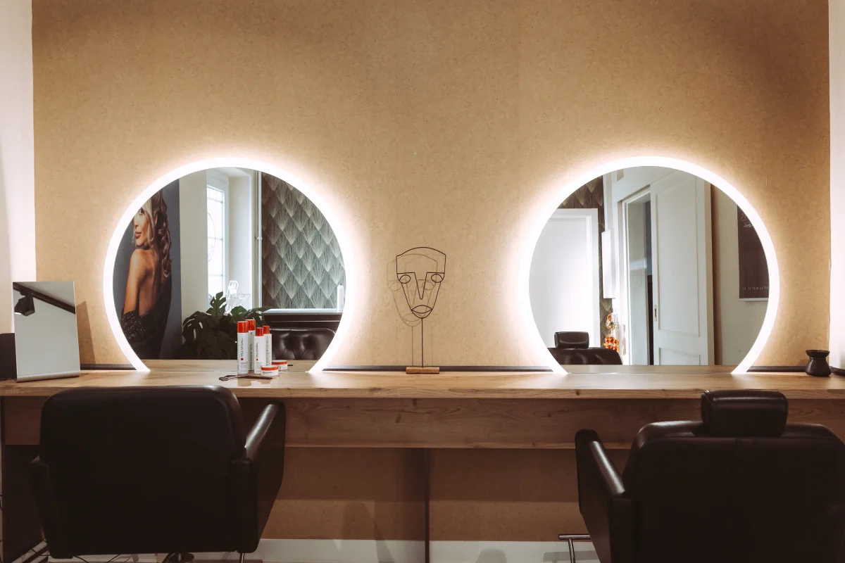 Professioneller Friseursalon mit zwei beleuchteten ovalen Spiegeln, Holzoberflächen und zwei schwarzen Ledersitzen, ideal für Imagefotos von Unternehmen.