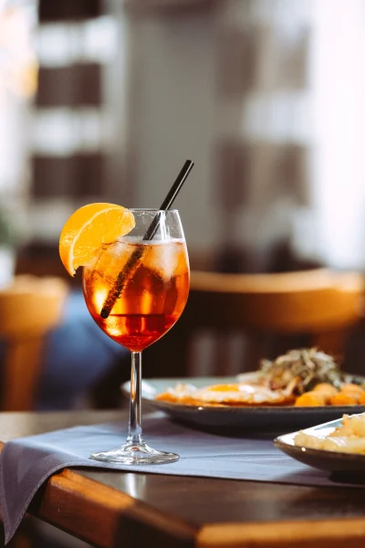 Ein erfrischender Aperol Spritz mit Eis und Orangenscheibe in einem Weinglas, serviert auf einem Tisch mit Essen im Hintergrund, ideal für Unternehmen Fotograf und Imagevideograf.