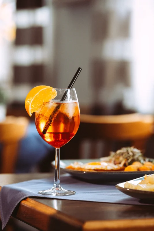 Ein erfrischender Aperol Spritz mit Eis und Orangenscheibe in einem Weinglas, serviert auf einem Tisch mit Essen im Hintergrund, ideal für Unternehmen Fotograf und Imagevideograf.
