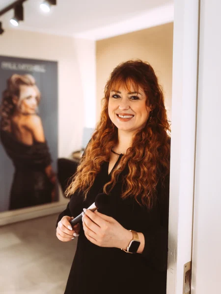 Ein professioneller Fotograf und Imagevideograf für Unternehmen, eine Frau mit roten lockigen Haaren und einem Lächeln, hält Make-up-Pinsel, im Hintergrund ein Poster mit einer Frau.