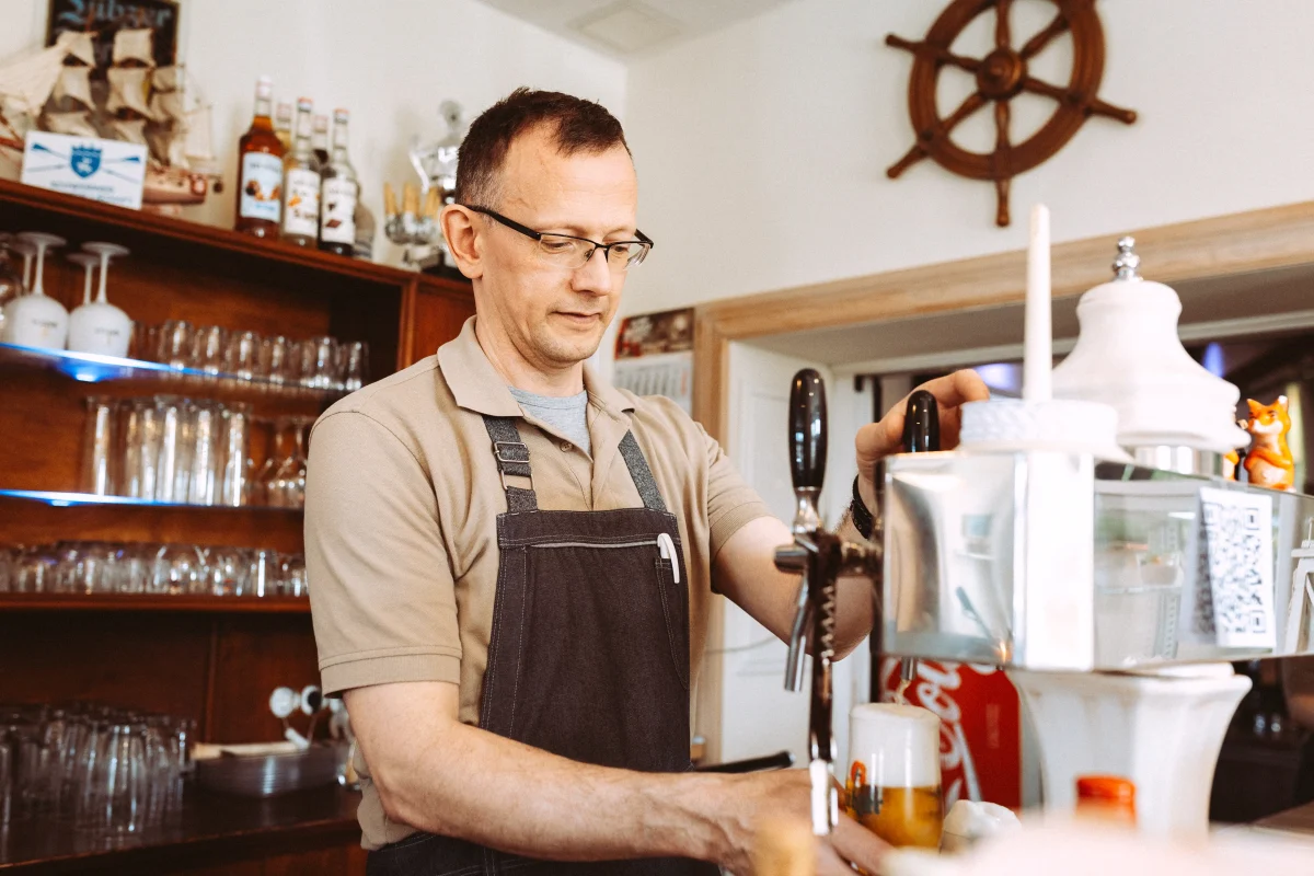 Ein Restraurantbesitzer zapft ein Bier in einer rustikalen Bar, mit Regalen voller Gläser und Flaschen im Hintergrund. Fotografiert von einem Businessfotografen.