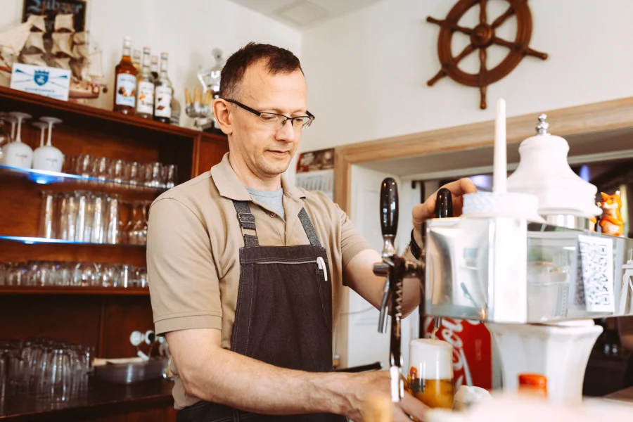 Ein Restraurantbesitzer zapft ein Bier in einer rustikalen Bar, mit Regalen voller Gläser und Flaschen im Hintergrund. Fotografiert von einem Businessfotografen.