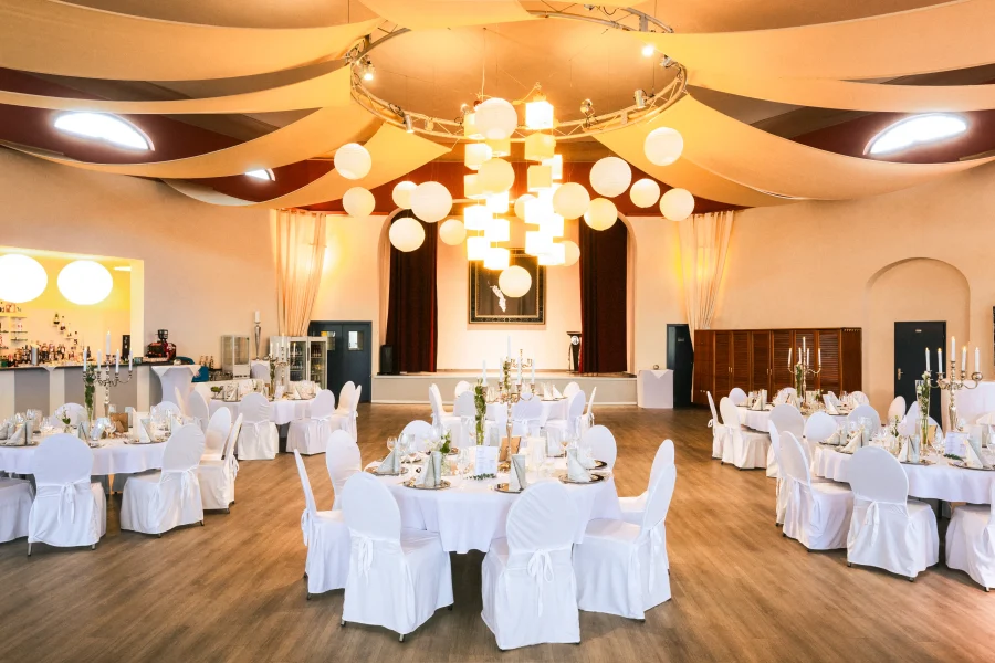 Festlich gedeckte Tische mit weißen Hussen und Blumenarrangements in einem eleganten Veranstaltungsraum mit indirekter Beleuchtung, bereit für eine Firmenfeier. Fotograf und Imagevideograf für Unternehmen.