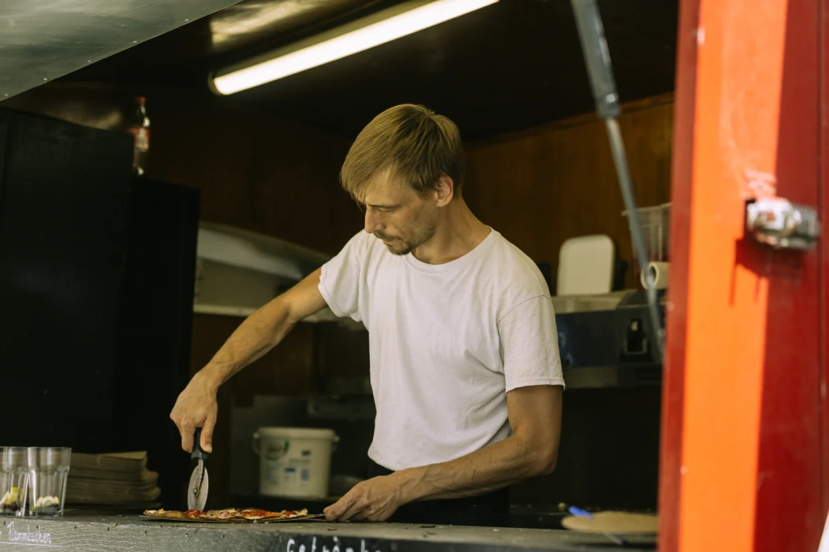 Ein konzentrierter Koch schneidet in einem Foodtruck einen Flammkuchen mit einem Pizzaschneider. Als erfahrener Unternehmen Fotograf und Imagevideograf fange ich solche authentischen Arbeitsprozesse ein, um die Identität und Handwerkskunst Ihres Betriebes professionell zu präsentieren.