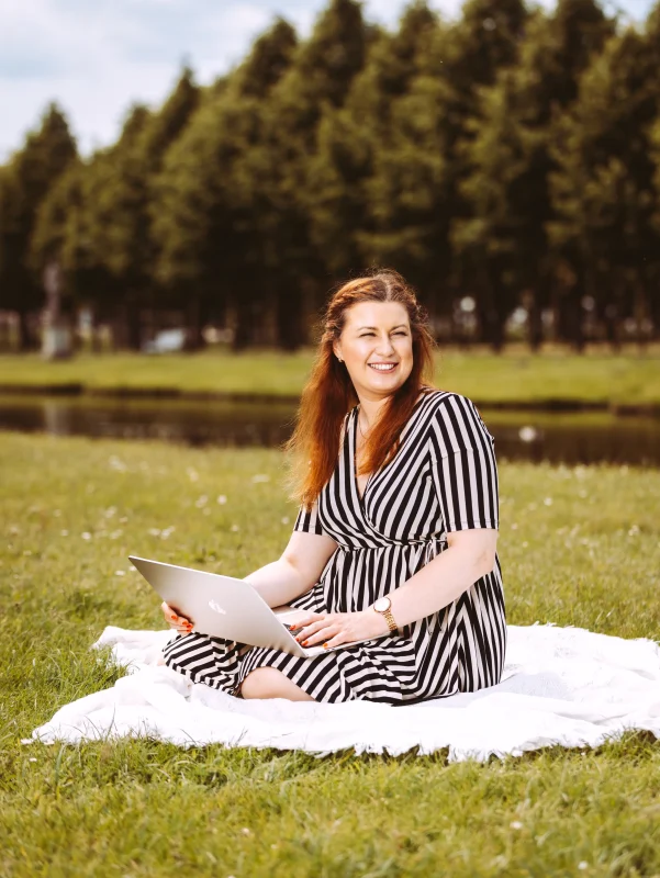 Frau im schwarzen Streifenkleid sitzt auf einer Decke im Park und arbeitet auf einem Laptop