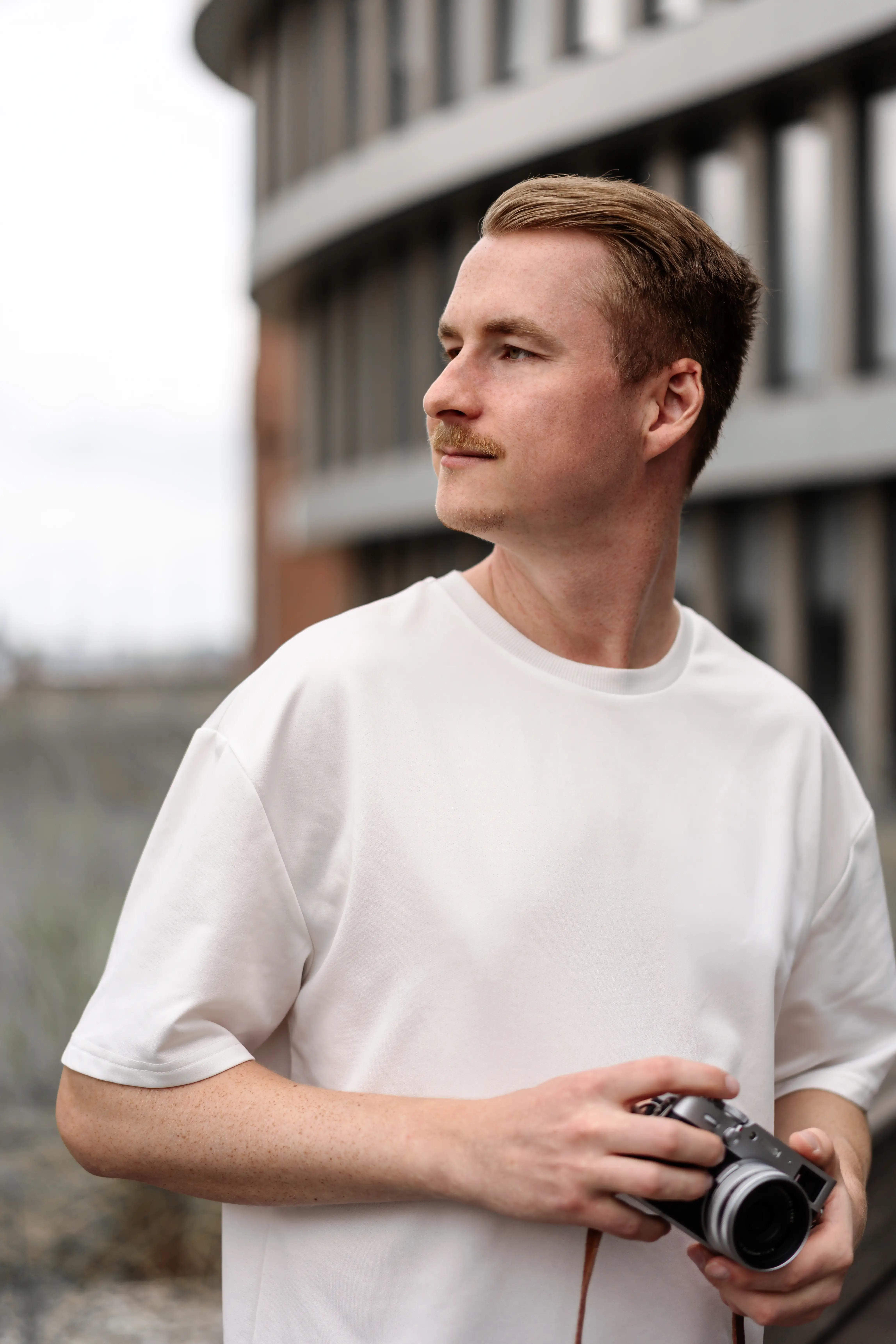 Ein professioneller Fotograf und Imagevideograf mit einer Kamera in der Hand, der vor einer modernen Unternehmensfassade in die Ferne blickt. Er trägt ein schlichtes weißes T-Shirt und steht für hochwertige Imagefotos und Imagevideos bereit.