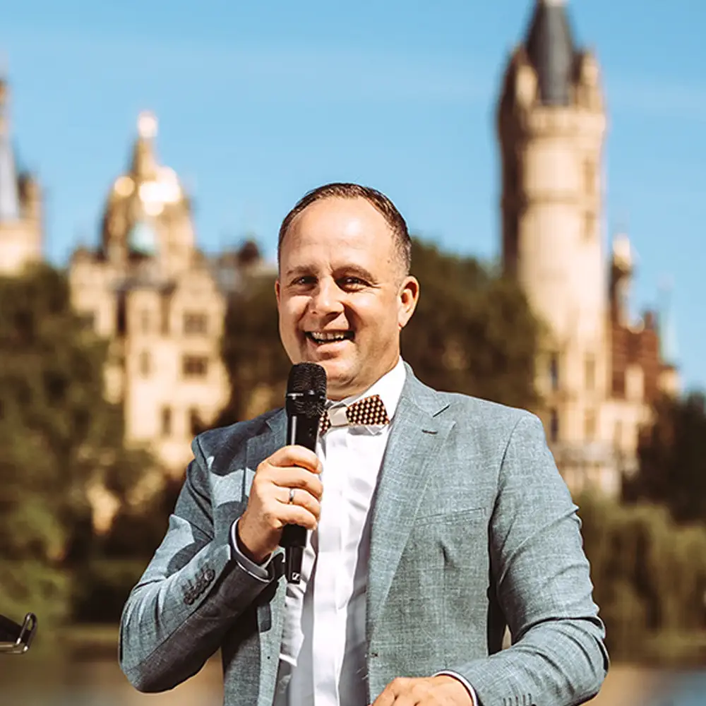 Ein professioneller Fotograf und Imagevideograf lächelt in ein Mikrofon, während er vor der malerischen Kulisse des Schweriner Schlosses spricht. Er trägt ein elegantes graues Sakko mit Fliege und präsentiert sich als kompetenter Partner für hochwertige Unternehmens-Medieninhalte. Ein professioneller Fotograf und Imagevideograf lächelt in ein Mikrofon, während er vor der malerischen Kulisse des Schweriner Schlosses spricht. Er trägt ein elegantes graues Sakko mit Fliege und präsentiert sich als kompetenter Partner für hochwertige Unternehmens-Medieninhalte.