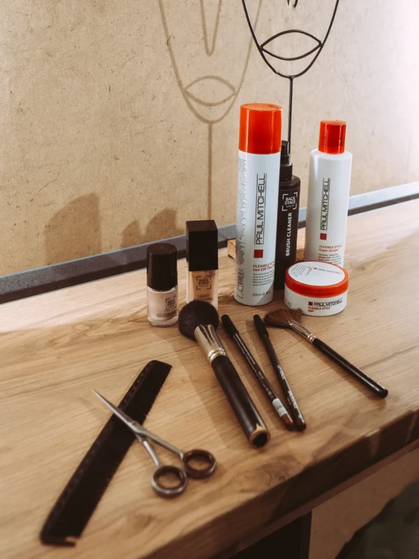 Professionelles Equipment für Friseure und Visagisten auf einem Holztisch: Paul Mitchell Haarpflegeprodukte, Make-up-Pinsel, eine Schere und ein Kamm vor einer minimalistischen Wandzeichnung. Als Unternehmen Fotograf und Imagevideograf setze ich Arbeitsutensilien ästhetisch in Szene.