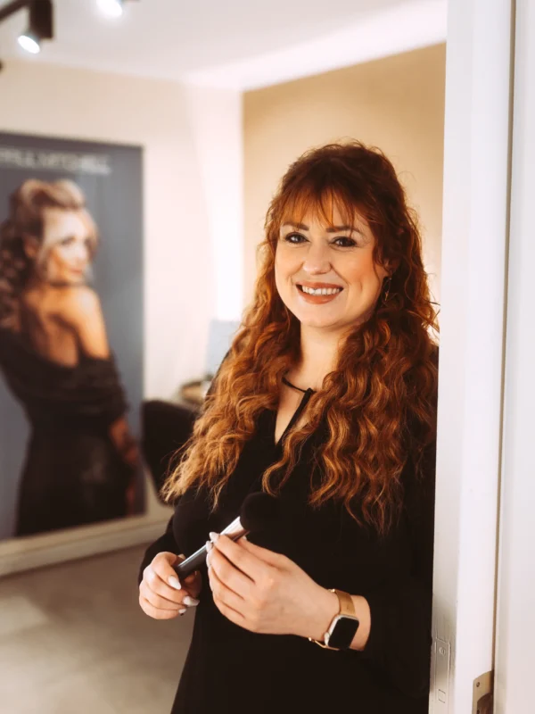 Eine professionelle Visagistin mit langem, rotbraunem Haar lächelt freundlich in die Kamera, während sie einen Make-up-Pinsel in den Händen hält. Im Hintergrund ist ein Poster eines Unternehmens-Fotografen und Imagevideografen zu sehen, das eine elegante Frau zeigt.