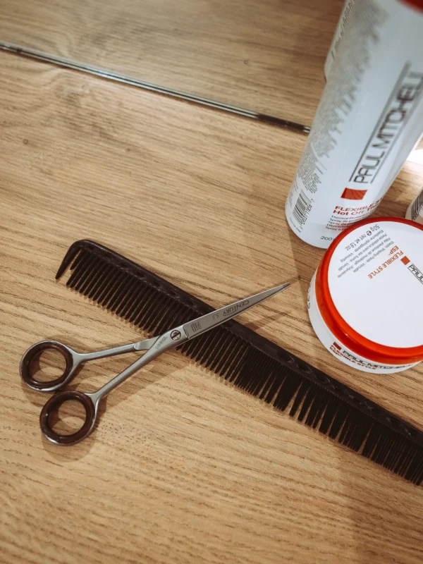 Friseurbedarf auf einem Holztisch: Eine Tondeo Century 5.5 Haarschere liegt auf einem schwarzen Kamm neben Paul Mitchell Styling-Produkten. Als professioneller Fotograf und Imagevideograf für Unternehmen setze ich hochwertige Arbeitsutensilien ästhetisch in Szene.