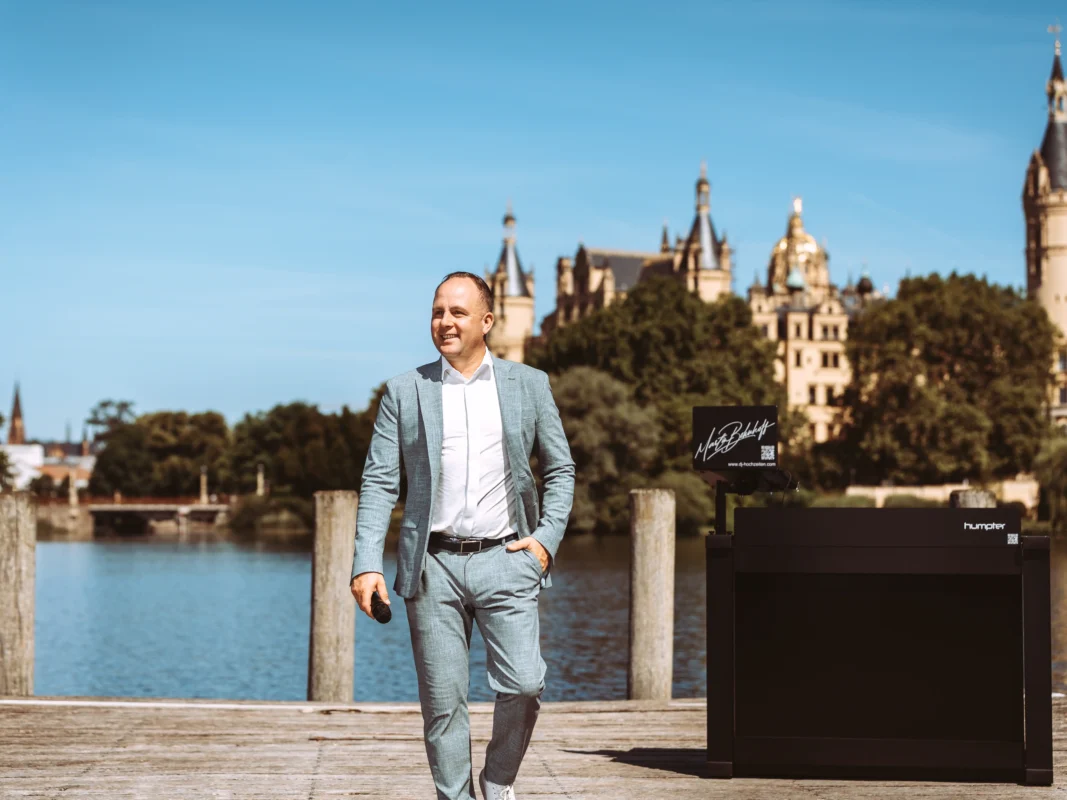 Ein professioneller Unternehmens-Fotograf und Imagevideograf in einem hellen Anzug lächelt auf einem Holzsteg vor dem Schweriner Schloss. Neben ihm steht ein mobiles DJ-Pult mit dem Schriftzug „Martin Behaff“. Die Szene strahlt Eleganz und Professionalität für Business-Auftritte aus.