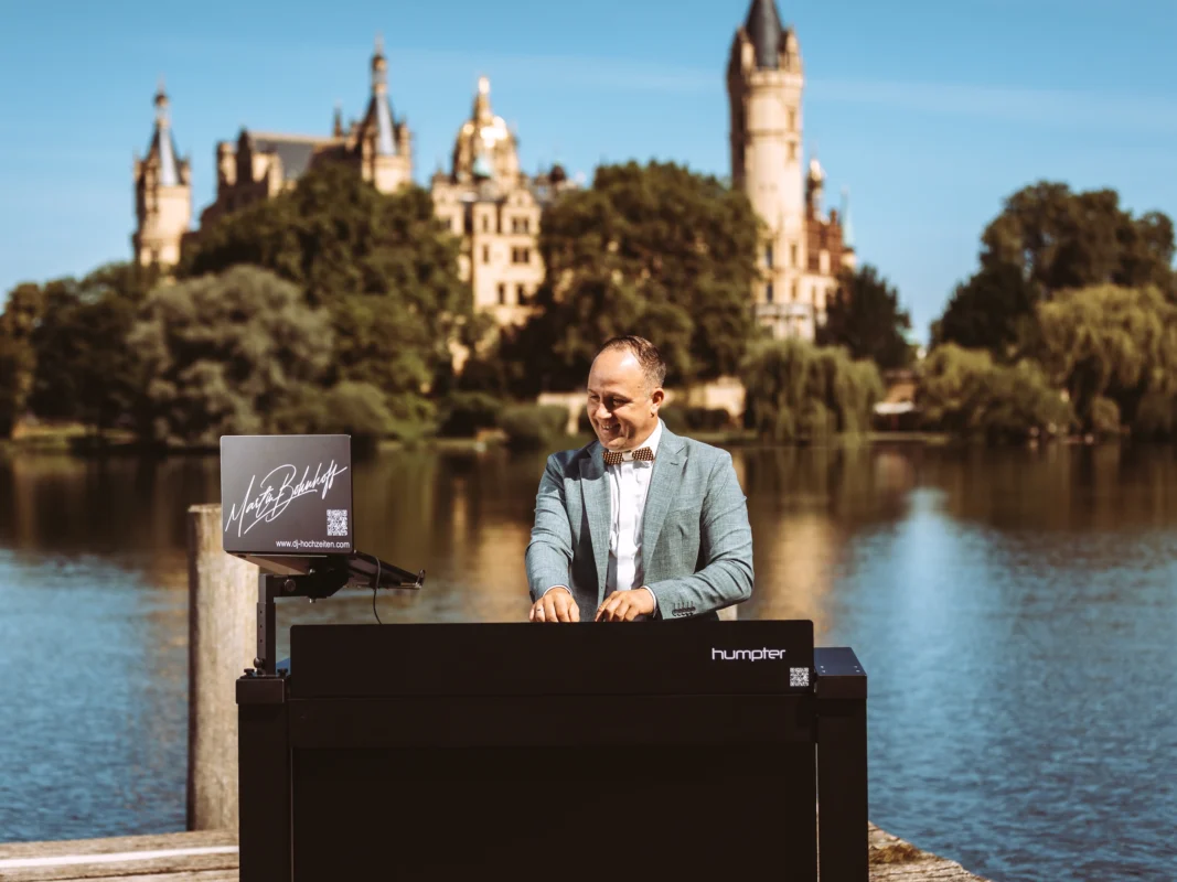 Ein professioneller DJ und Musiker in einem hellblauen Sakko mit Fliege steht hinter einem Humpter-DJ-Pult vor dem malerischen Schweriner Schloss. Als erfahrener Unternehmen Fotograf und Imagevideograf halte ich solche hochwertigen Business-Momente für Sie fest.