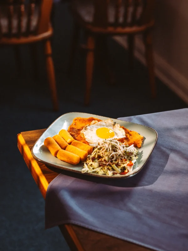 Ein appetitlich angerichtetes Wiener Schnitzel mit Spiegelei, Kroketten und frischem Salat auf einem quadratischen Teller, fotografiert in einem Restaurant. Professionelle Food-Aufnahme, erstellt von einem Unternehmen Fotograf und Imagevideograf für hochwertige visuelle Inhalte.