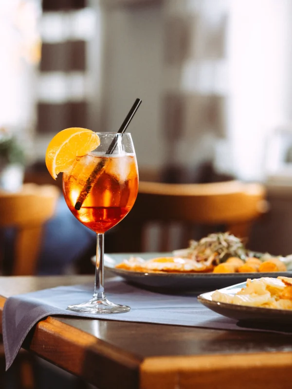 Ein erfrischender Aperol Spritz mit einer Orangenscheibe steht auf einem gedeckten Holztisch neben einem Teller mit Frühstück. Diese atmosphärische Aufnahme zeigt die Qualität, die ich als Unternehmen Fotograf und Imagevideograf für Ihre professionelle Bildsprache liefere.