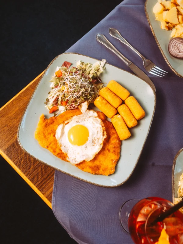 Ein appetitlich angerichtetes Schnitzel mit Spiegelei, Kroketten und frischem Salat auf einem hellblauen Teller, fotografiert von einem professionellen Unternehmen Fotograf und Imagevideograf für eine hochwertige kulinarische Präsentation.