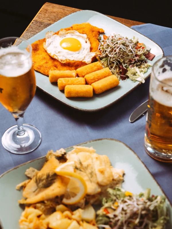 Zwei appetitliche Teller mit Schnitzel, Spiegelei, Kroketten und frischem Salat stehen neben zwei Biergläsern auf einem blauen Tischtuch. Als professioneller Unternehmen Fotograf und Imagevideograf fange ich solche kulinarischen Momente für die Gastronomie ein.