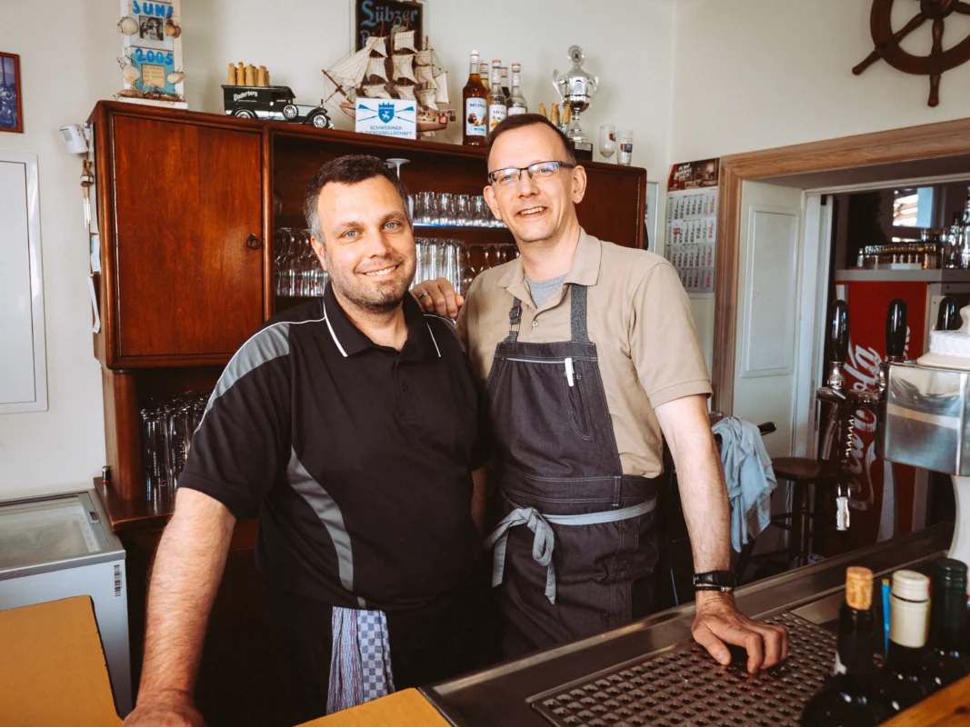Zwei freundliche Gastronomen hinter einem Tresen in einer Bar, fotografiert von einem professionellen Unternehmen Fotograf und Imagevideograf. Sie tragen Arbeitskleidung und lächeln in die Kamera, im Hintergrund stehen ein Modellschiff und eine Vitrine mit Gläsern.